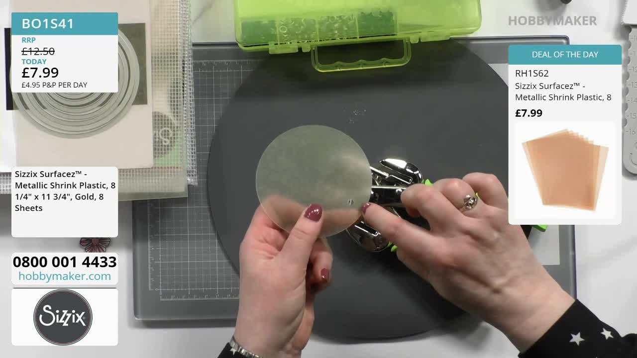 A hand presses a punch through a circular piece of gold shrink plastic. The tool, a Sizzix Surfacez, makes a clean hole.