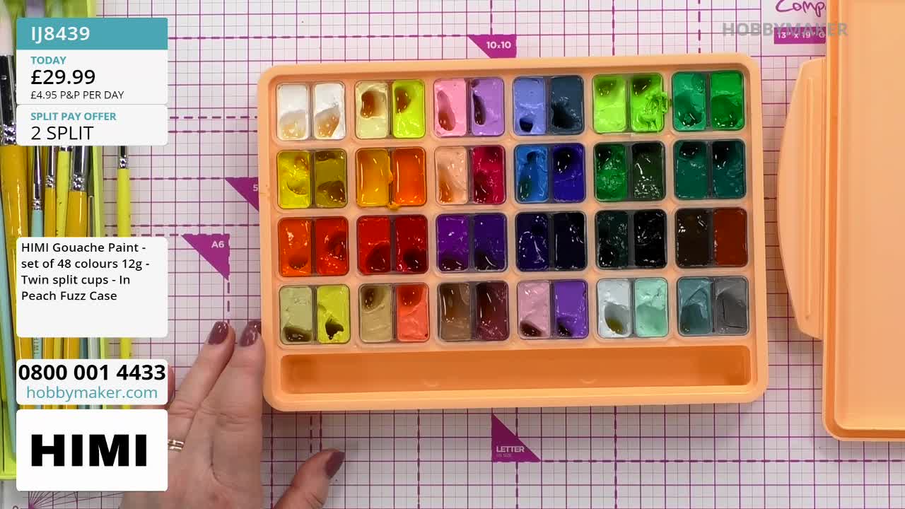 A hand reaches towards a tray filled with 48 small pots of gouache paint, each a different vibrant colour. The tray, a soft peach colour, sits on a grid mat, and a set of paintbrushes is visible to the left.