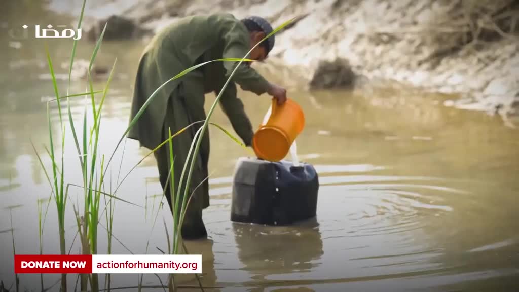 A man in a green tunic carefully pours water from an orange bucket into a black container submerged in a muddy stream. Tall green reeds stand to his left, their reflections shimmering on the water's surface.