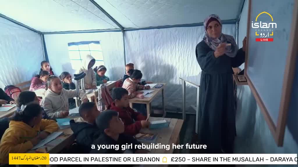 A woman stands at a whiteboard, addressing a classroom of children seated at desks. The children, some wearing hoodies, are focused on the lesson.