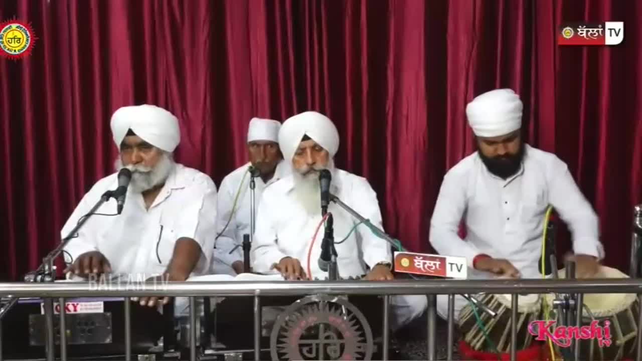 The man on the right plays a tabla with his hands, his fingers moving quickly across the drumheads. Beside him, a man in the center sings into a microphone, his white beard prominent.