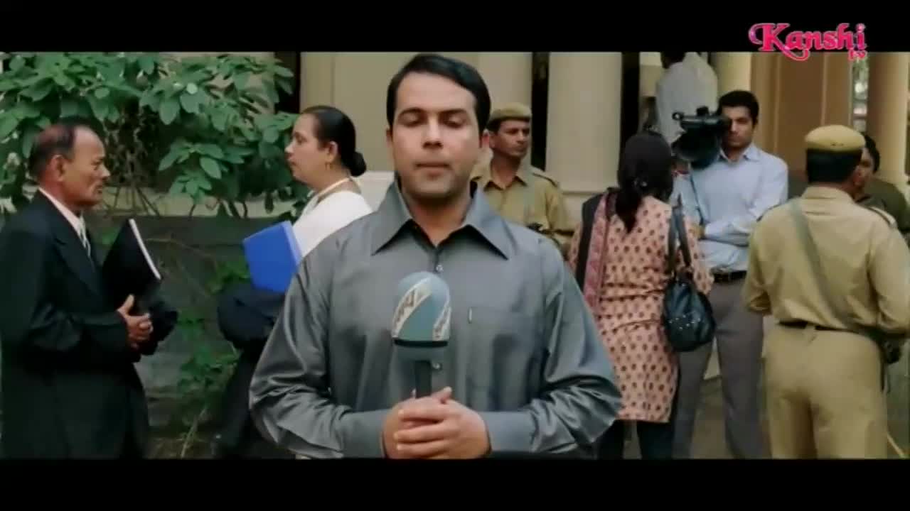 A reporter stands with a microphone, ready to speak for Kanshi TV. Behind him, a cameraman films as people mill about.