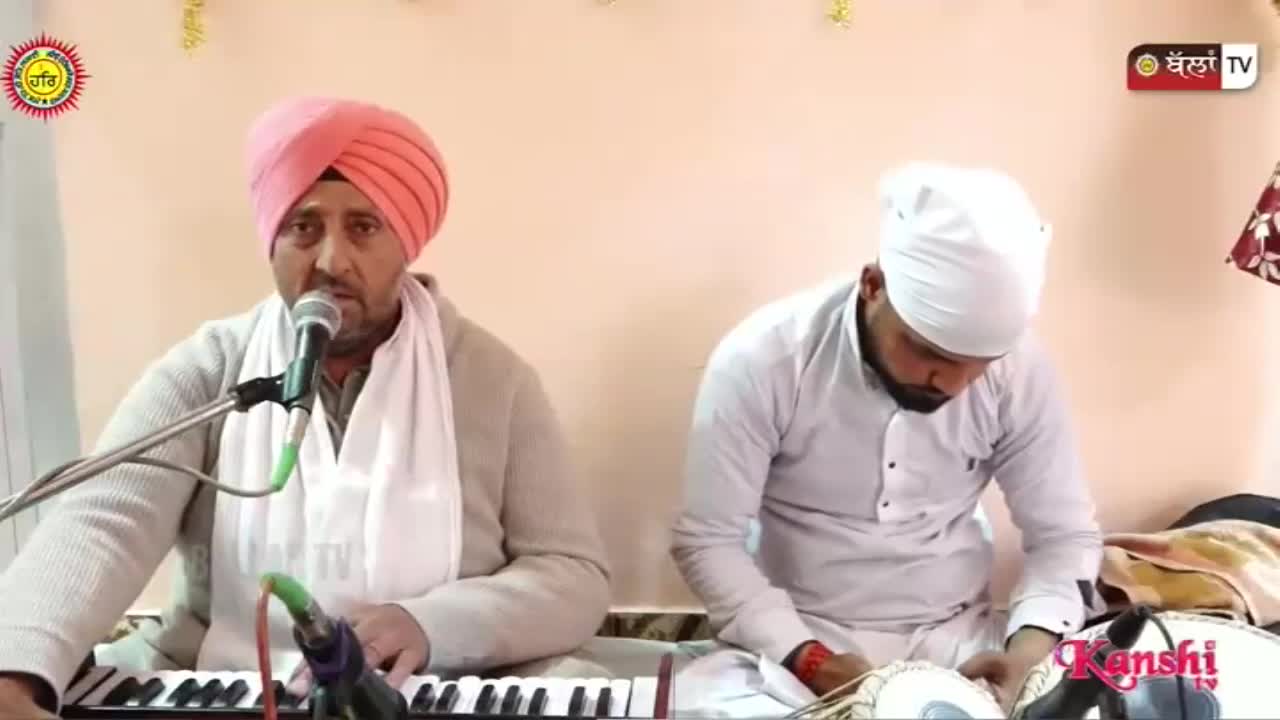 A man in a pink turban plays a keyboard while singing into a microphone. Beside him, another man in a white turban plays a tabla.