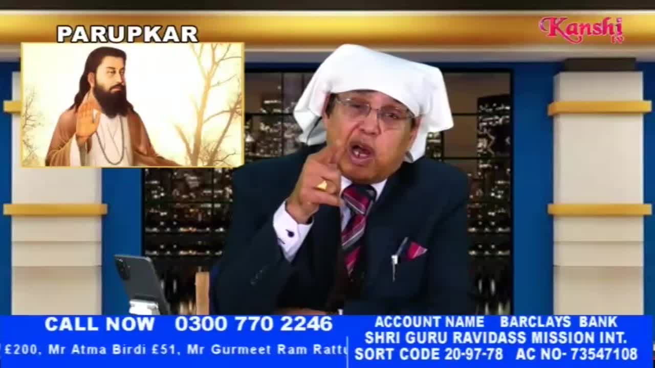 A man in a suit and white head covering speaks animatedly, gesturing with his right hand. On a screen behind him, a portrait of a man with a beard is displayed.