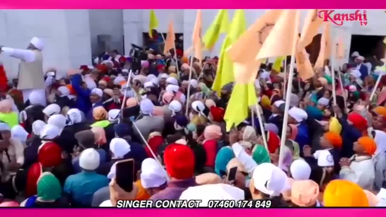 A man in a cream vest stands on a raised platform, pointing to a large crowd gathered below. Many people in the crowd are holding up colourful flags, some yellow and some orange, as they look towards the platform.