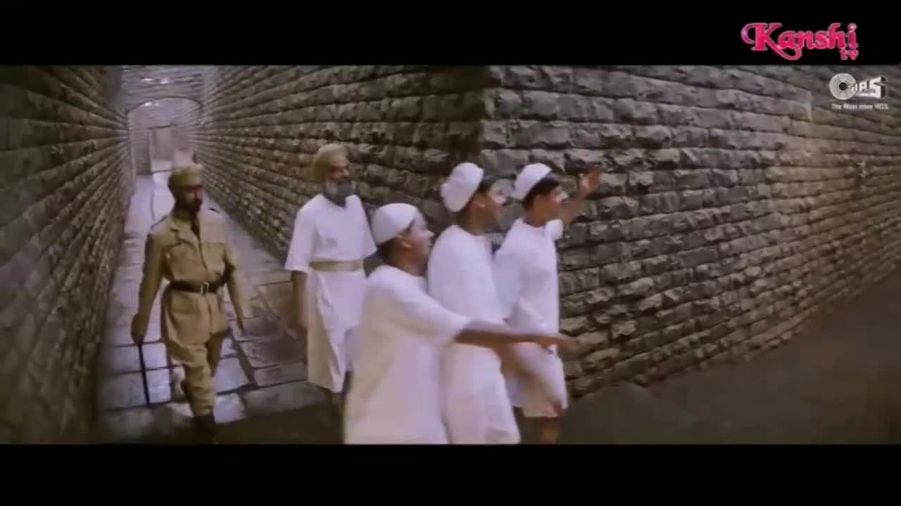A man in a khaki uniform walks ahead of a group of men in white robes. They are moving down a stone corridor, the Kanshi TV logo visible in the corner.