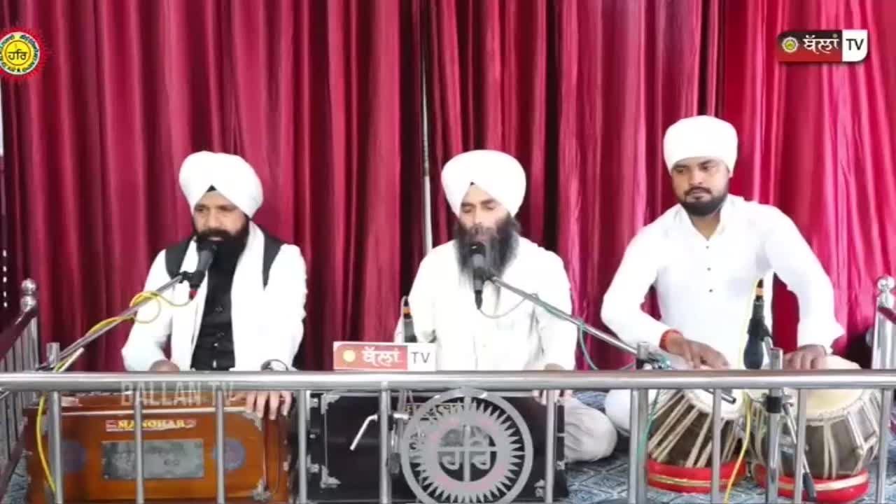 Three men in white turbans sit behind a metal railing. The man on the left plays a harmonium, while the man on the right taps out a rhythm on a tabla. The middle man sings into a microphone, his long beard reaching his chest.