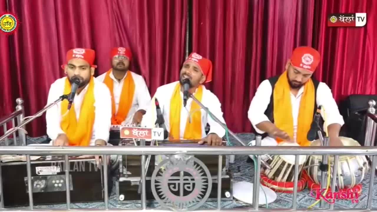 A man sings into a microphone, his voice amplified by the red curtains behind him. To his left, another man plays a keyboard, while to his right, a third man beats a tabla. The Kanshi TV logo is visible in the corner.