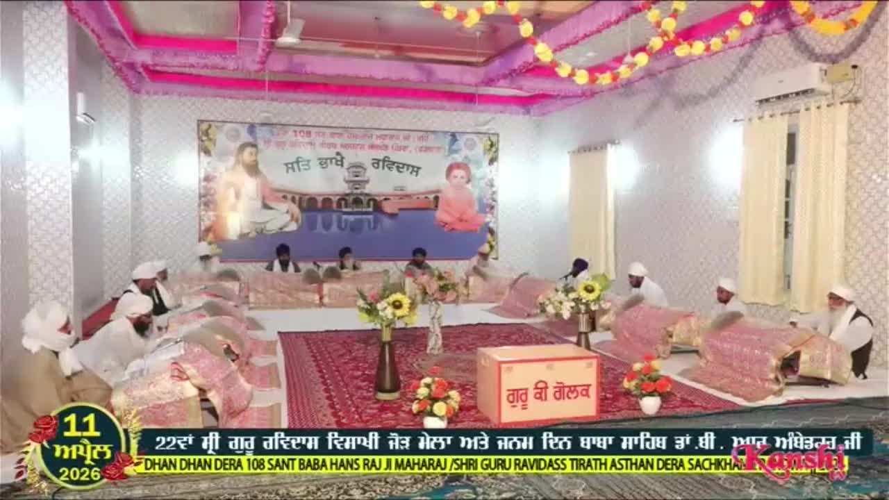 A group of men, dressed in white turbans and robes, are seated on the floor of a decorated hall. They are gathered around low tables covered with pink fabric, listening to someone speak. The scene is being broadcast live on Kanshi TV.