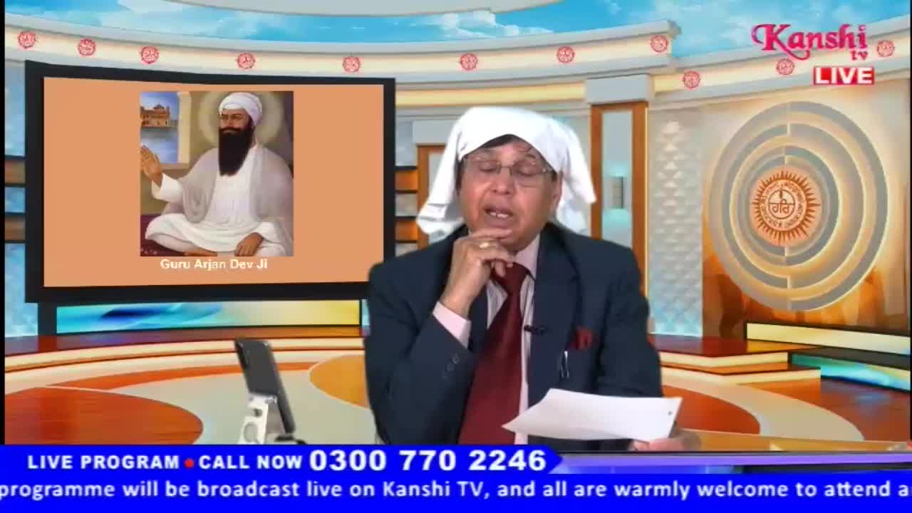 A man in a suit and a white cloth draped over his head speaks into a microphone. Behind him, a large screen displays a portrait of Guru Arjan Dev Ji.