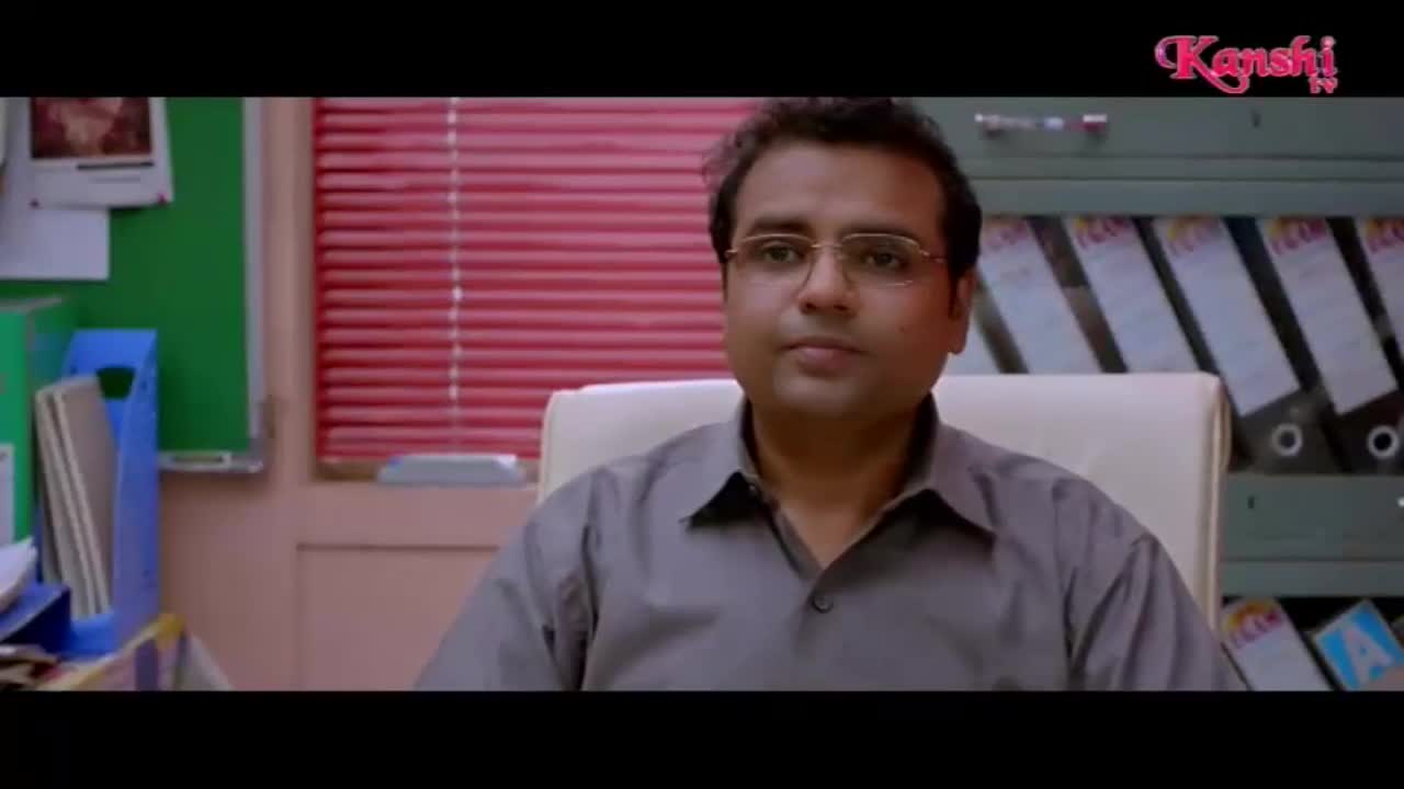 A man in glasses sits in an office chair, looking directly ahead. Behind him, red blinds cover a window, and shelves are filled with ring binders.