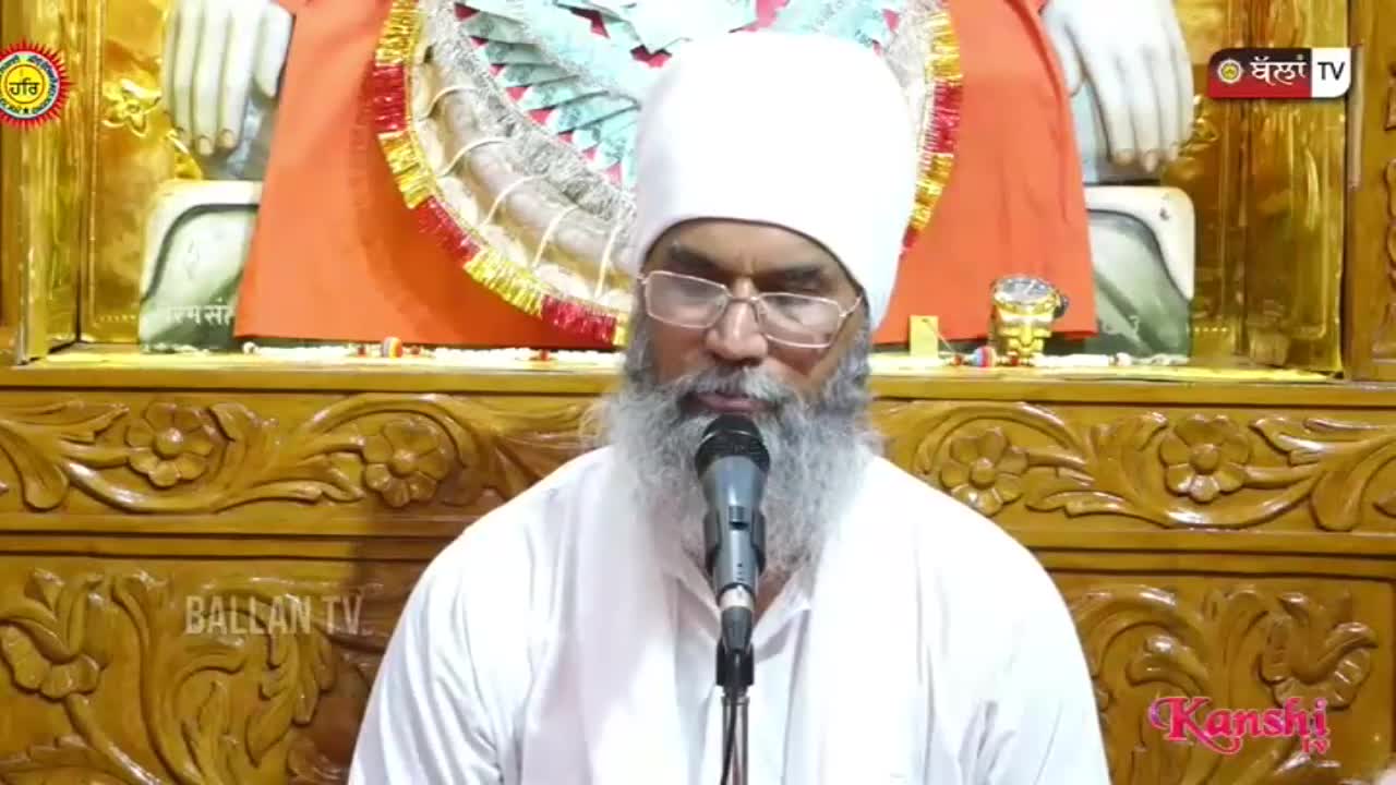 A man in a white turban and glasses speaks into a microphone. The background features ornate wooden carvings and a logo for Kanshi TV.