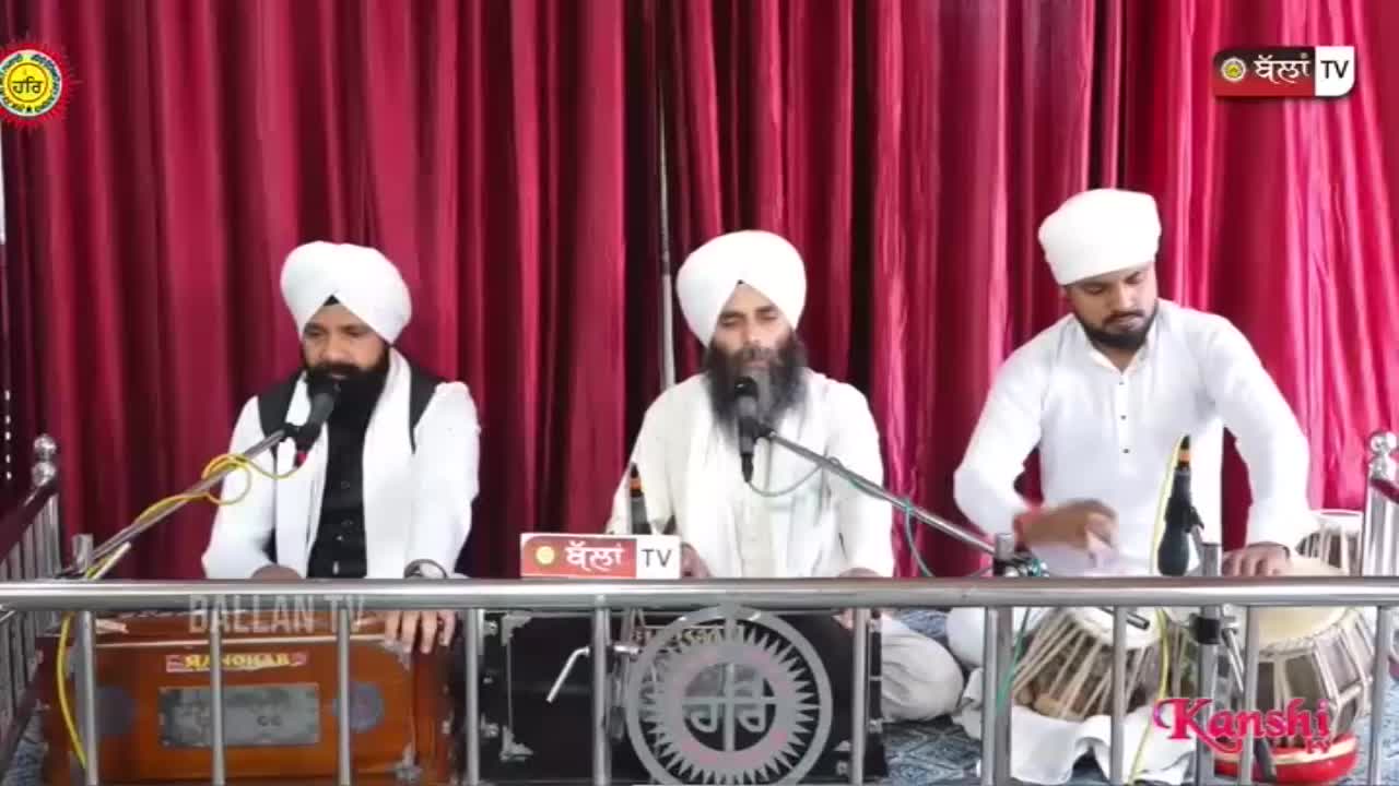 Three men in white turbans are seated, singing into microphones. One man plays a harmonium, another a tabla, as a Kanshi TV broadcast unfolds.