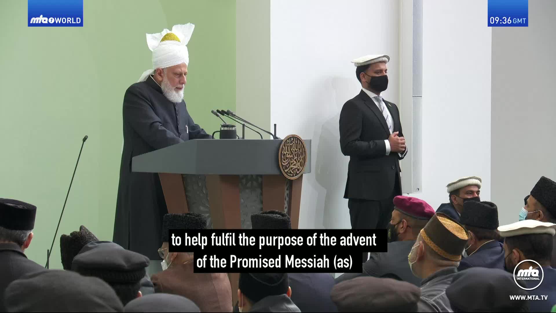 A man in a black suit and a white cap stands attentively beside a speaker at a podium. The speaker, also in a dark robe and white turban, addresses an audience seated before them.