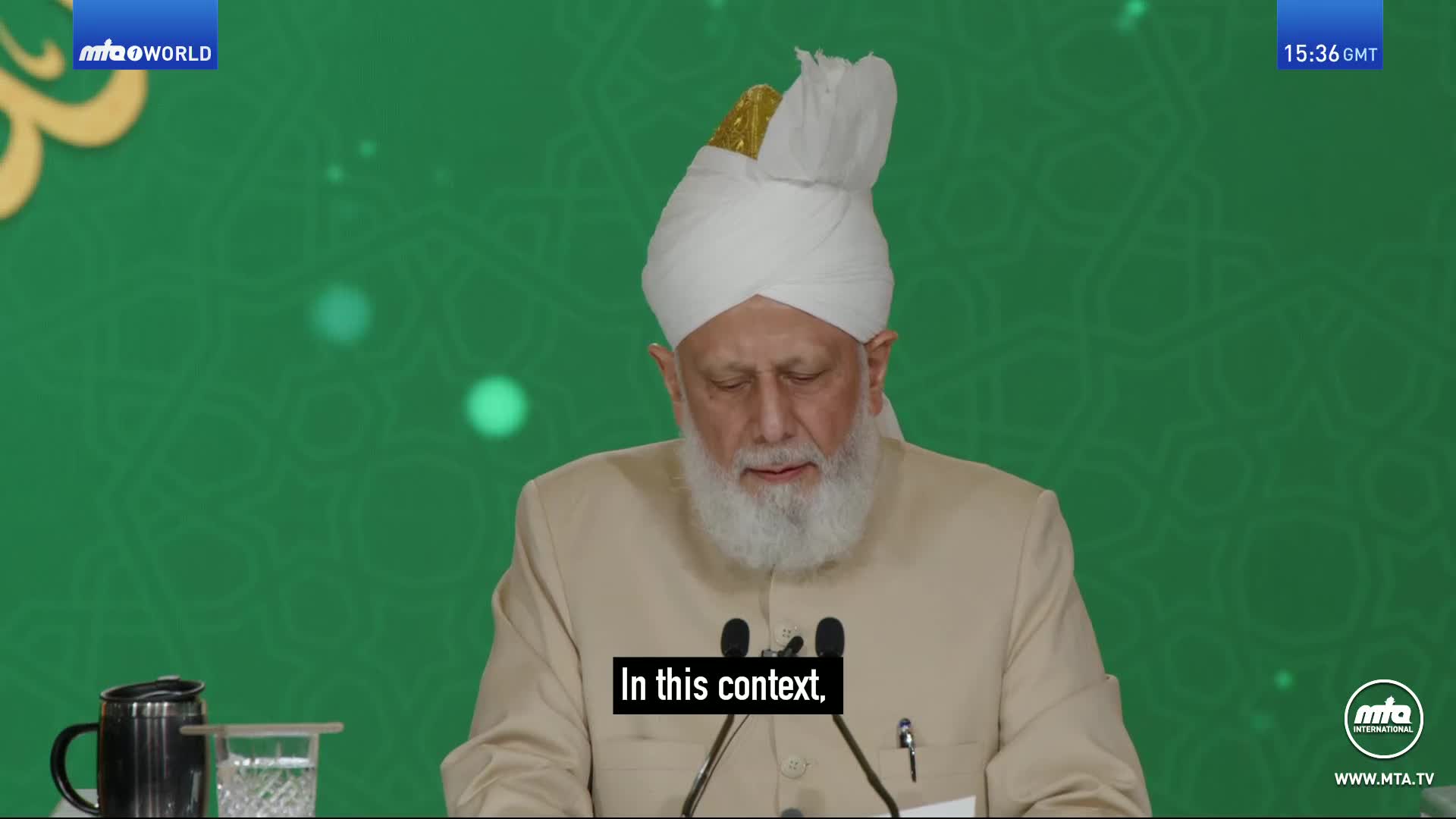 A man in a turban and light-colored jacket speaks into a microphone, his gaze lowered. A dark mug and a glass of water sit on the table before him, with the MTA World logo visible in the corner.