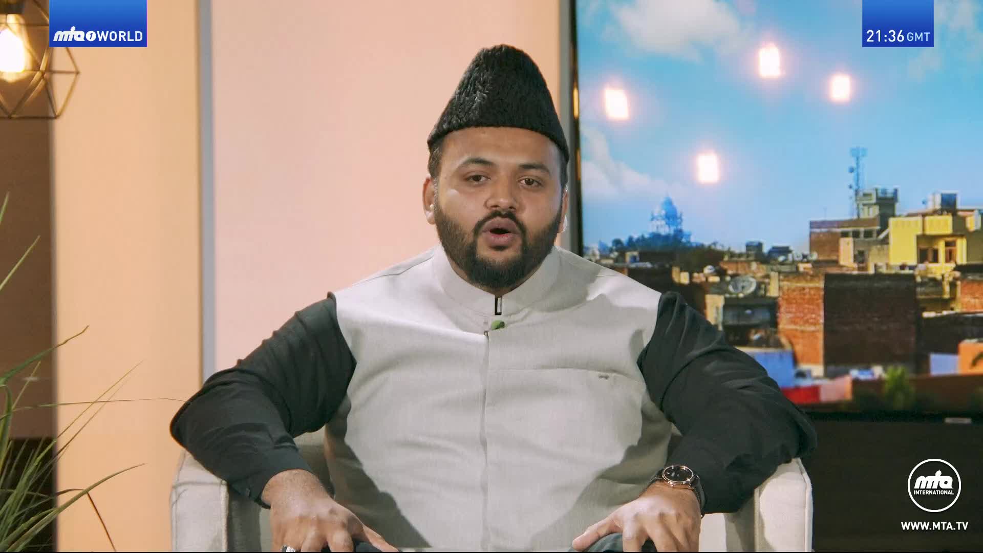 A man in a black cap and grey vest speaks directly to the camera. Behind him, a screen displays a cityscape with a clock showing 21:36 GMT.