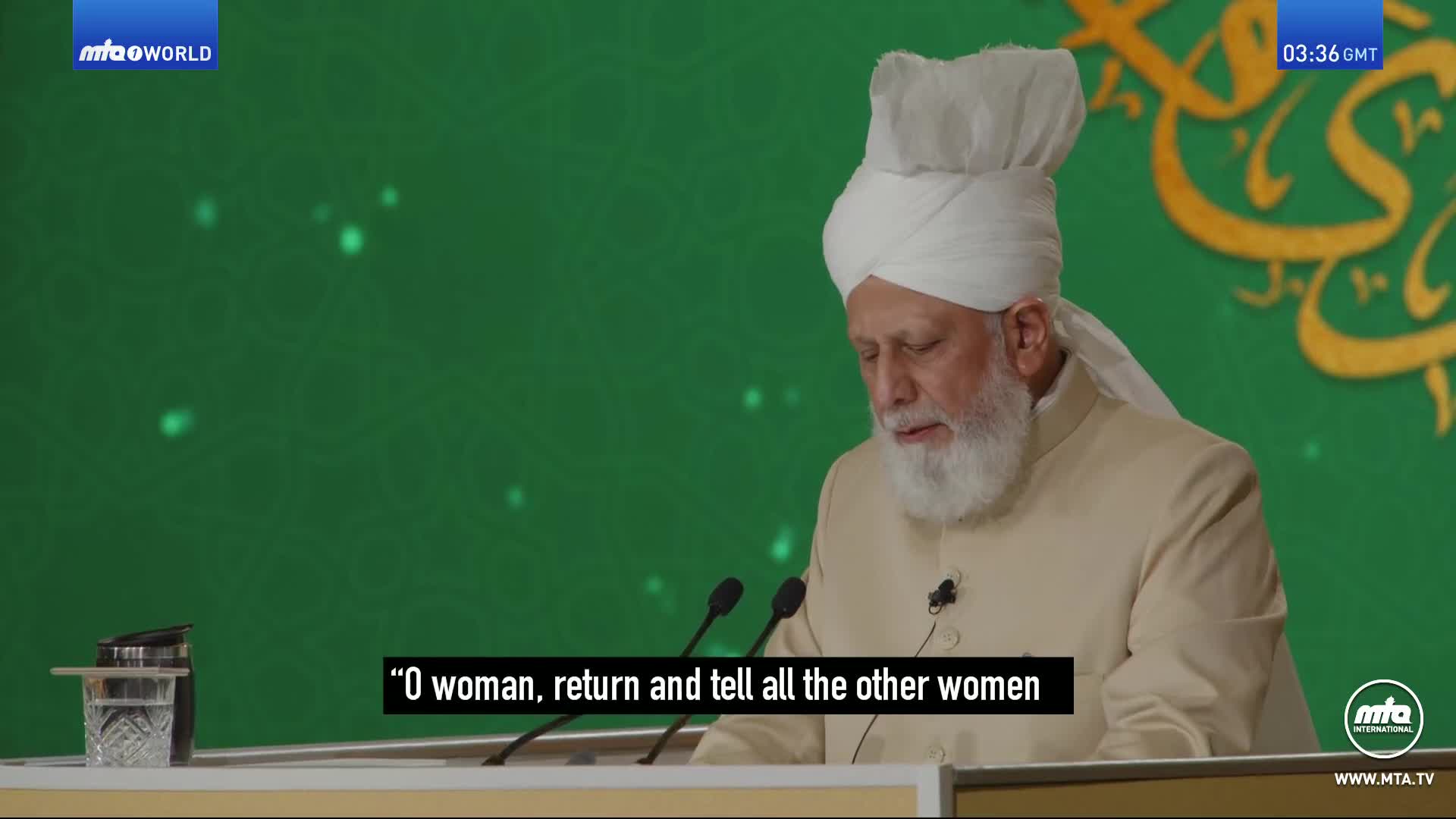 A man with a white turban and beard speaks at a podium. The background is a bright green screen with some glowing orbs and Arabic script.