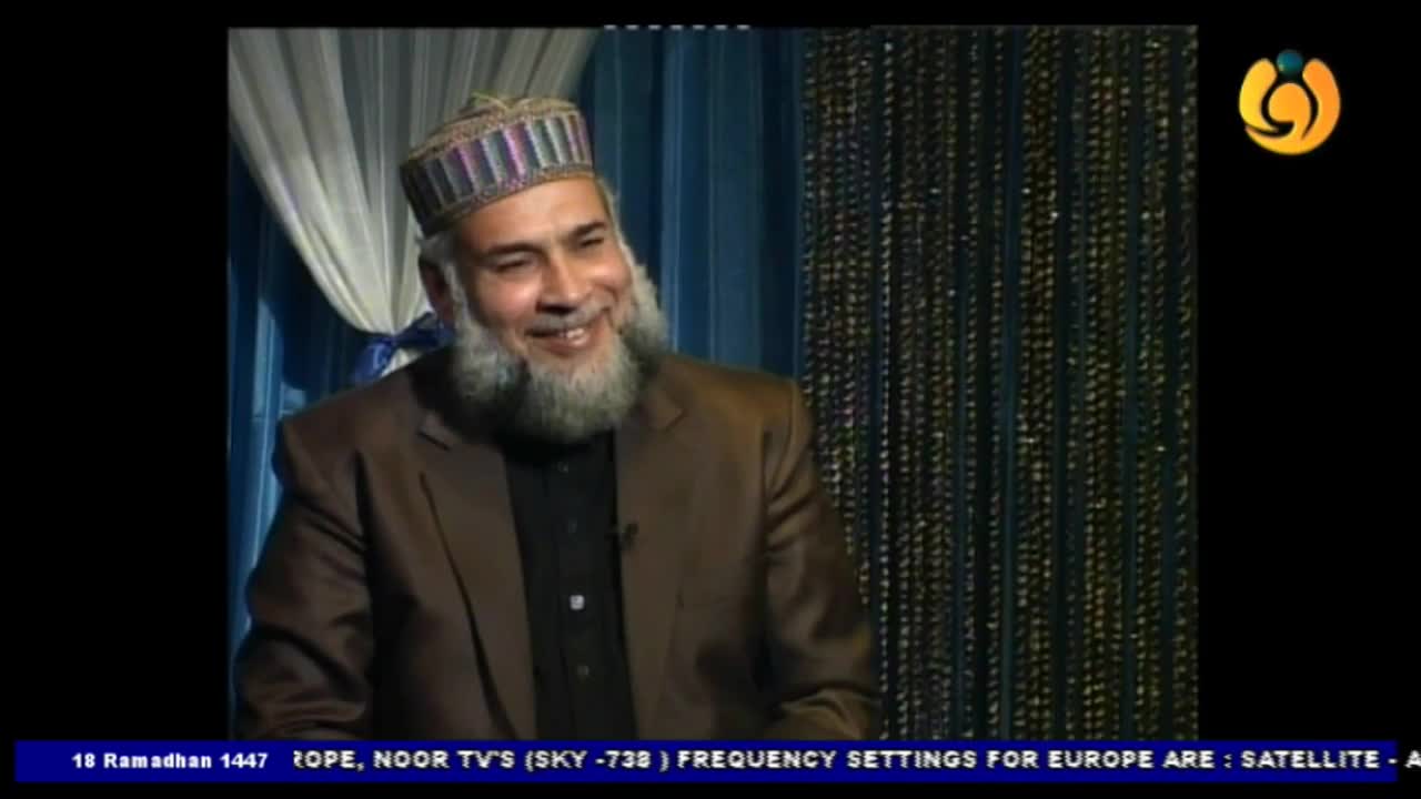 A man with a full beard and a patterned cap smiles warmly, his eyes crinkling. He wears a brown suit jacket over a dark shirt, seated against a backdrop of draped fabric.