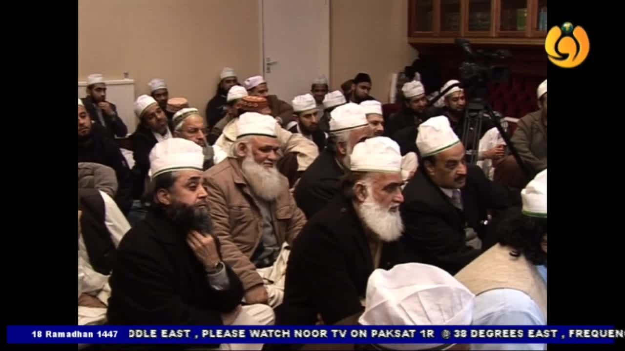 A group of men, many with white turbans and beards, are seated in a room. A camera operator films them from the side.