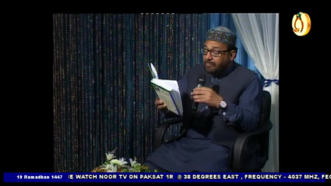 A man in a blue outfit and a cap reads from a book, holding a microphone in his other hand. The backdrop behind him is a dark curtain with shimmering vertical threads.