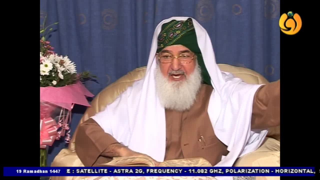 A man in a green turban and white head covering speaks animatedly, gesturing with his right hand. He's seated in a plush chair, with a bouquet of flowers to his left.