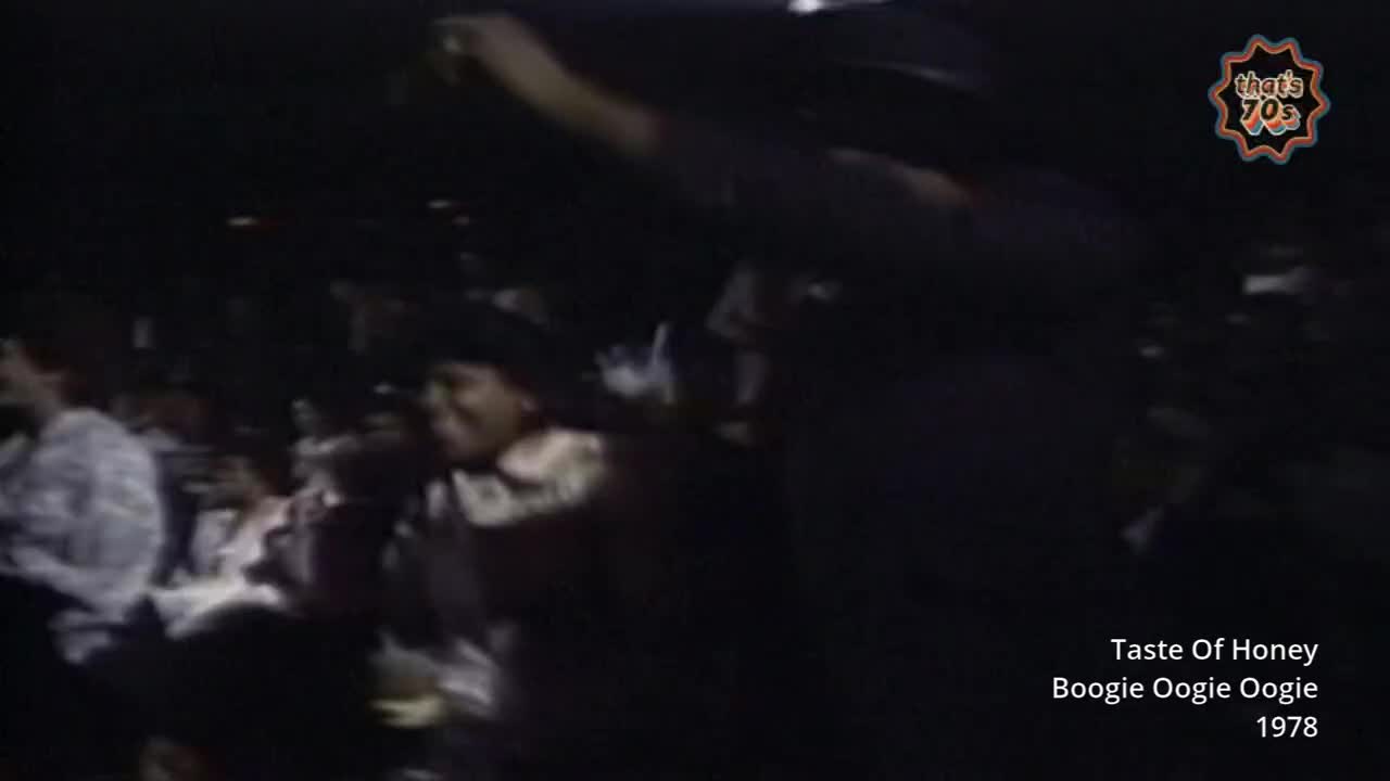 A crowd is packed together, faces turned upwards, some with hands raised. The air is thick with anticipation as the music plays.