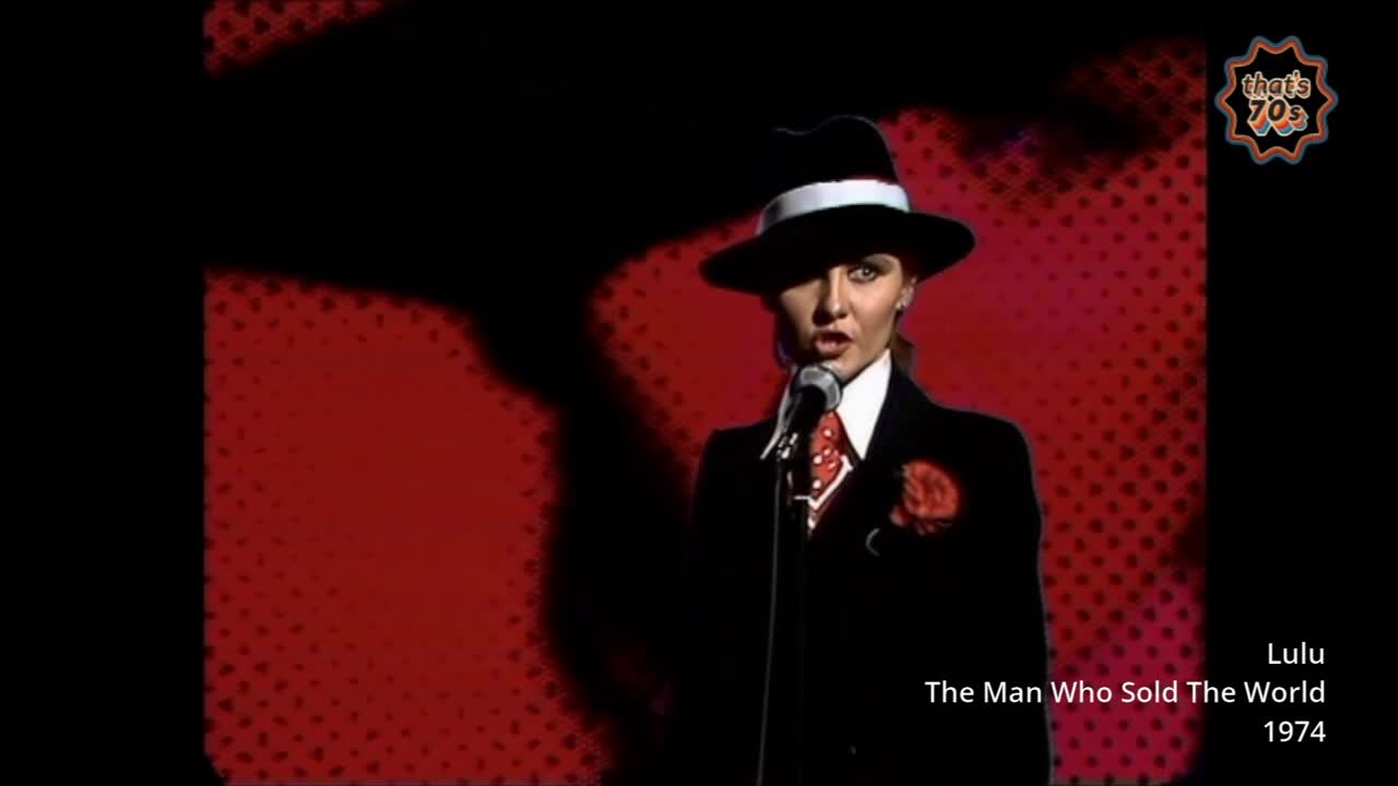 A woman in a sharp suit and fedora stands at a microphone, singing. The stage behind her is a deep red with a subtle dot pattern.