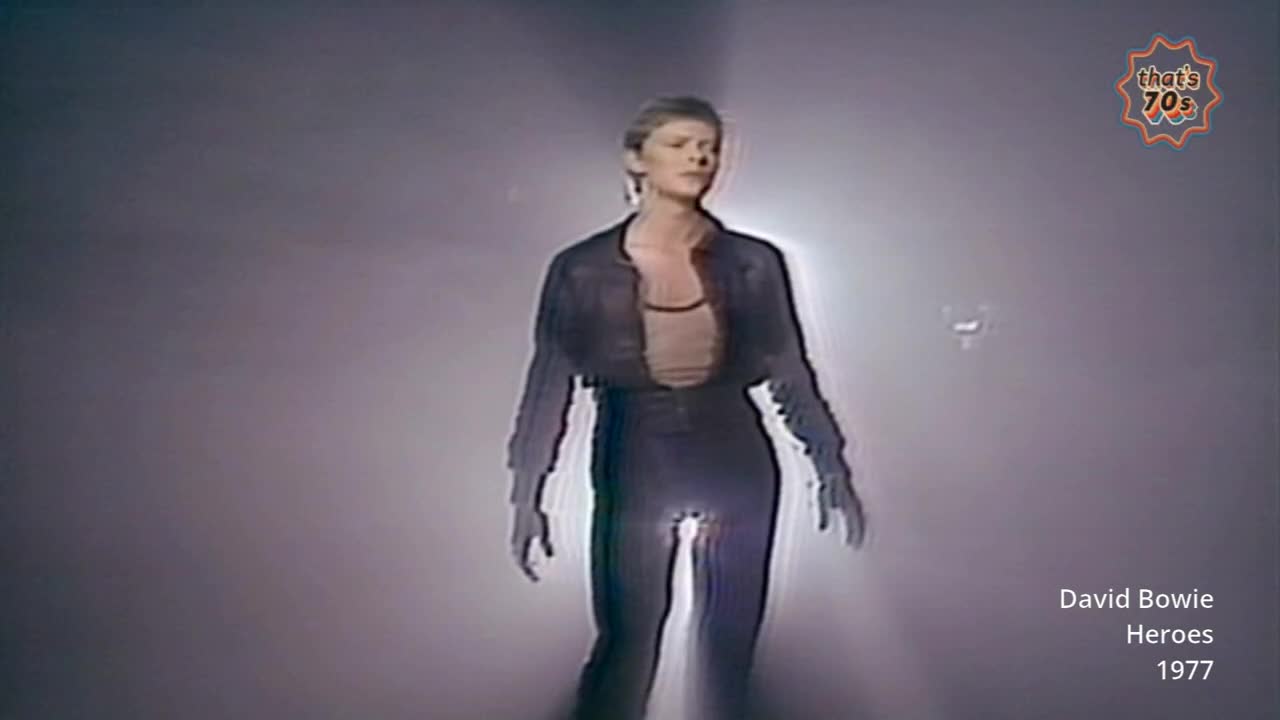 David Bowie stands under a bright spotlight, his dark jacket and trousers catching the light. The 1977 performance captures a moment of iconic British music history.