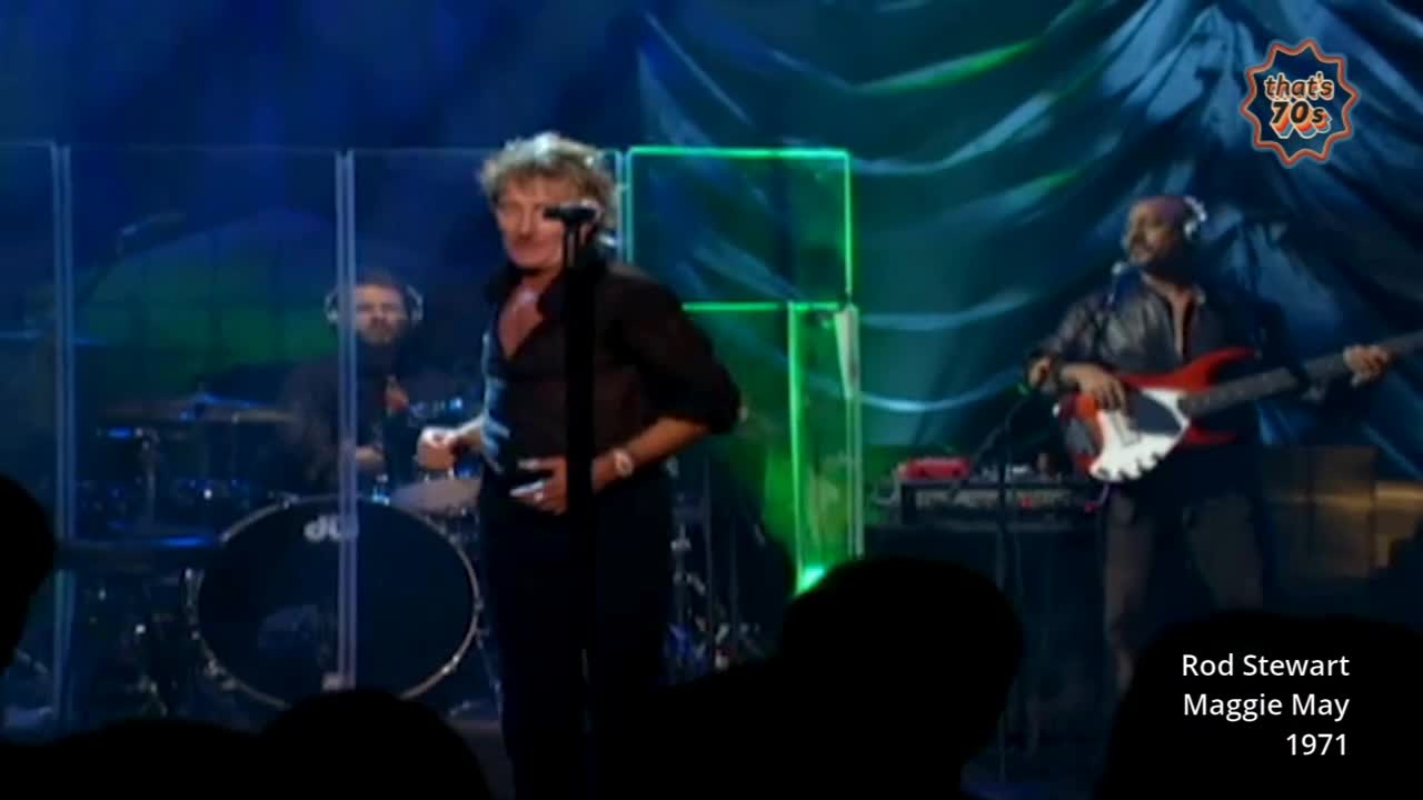 Rod Stewart is on stage, singing into a microphone. A bassist plays a red electric guitar to his right, while a drummer is visible behind him.