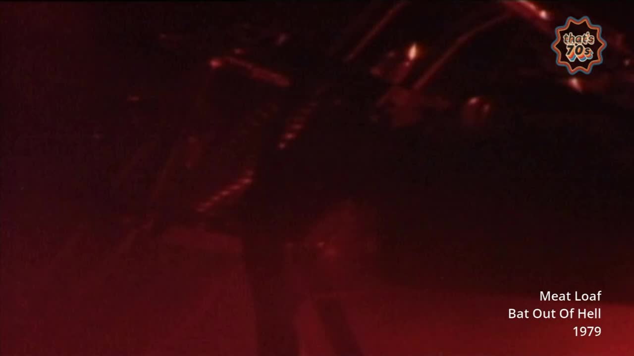 The stage lights cast a deep red glow, illuminating a figure in a dark costume. A chain-link fence, or something similar, hangs above, catching the light.