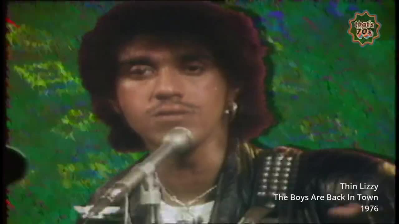 A man with a dark curly wig and a mustache sings into a microphone, his leather jacket studded with metal. The backdrop is a swirling green and yellow pattern, typical of a 1970s television broadcast.