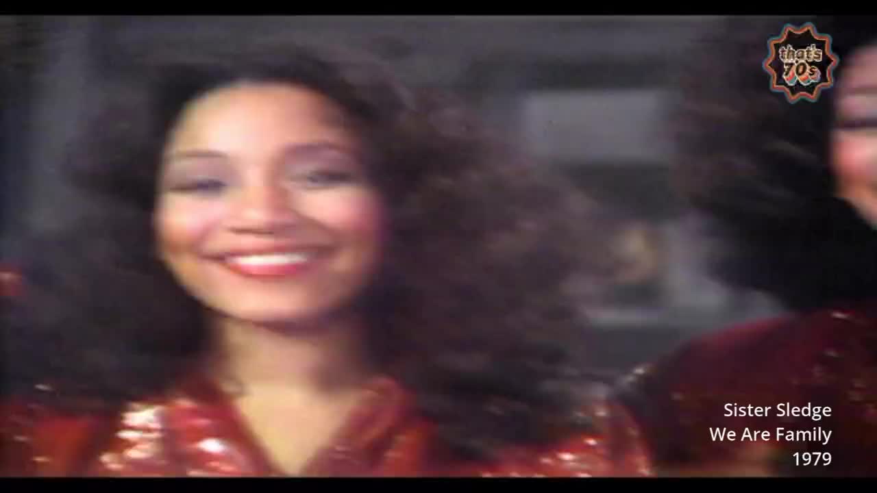 A woman with dark, curly hair smiles broadly, her red sequined top catching the light. Another woman, also in sequins, is visible to her right, her face turned away.