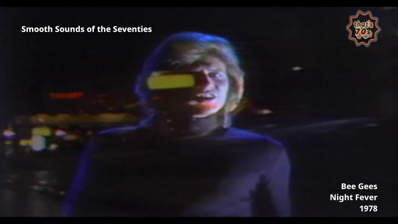 A man with a bright yellow visor over his eyes stands in the dark, his face illuminated by the lights behind him. The backdrop is a blurry cityscape, suggesting a night out in Britain during the late seventies.