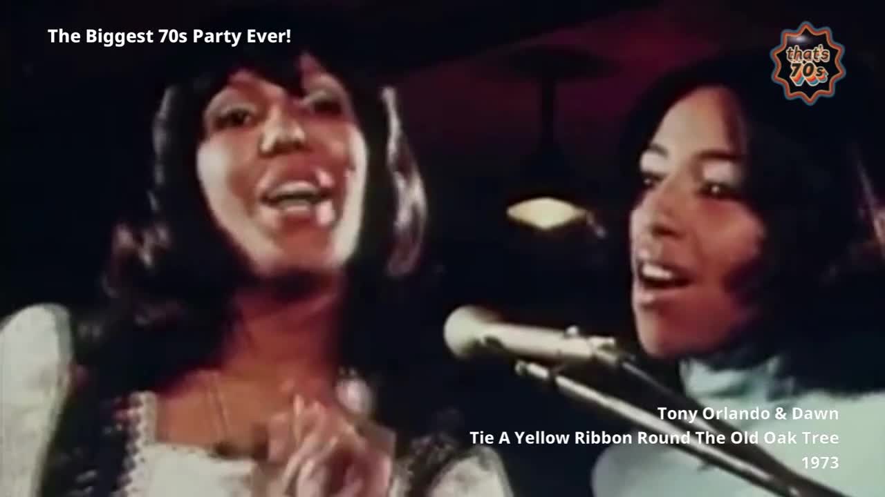 Two women sing into microphones, their faces illuminated by stage lights. The woman on the left, with dark, wavy hair, wears a white, ruffled top. The woman on the right, with straight dark hair, has a light blue collar visible.