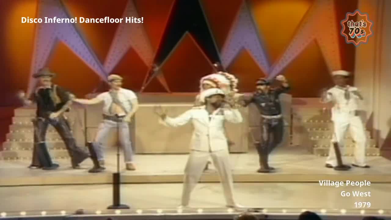 A group of men in distinct costumes are performing on stage. One man in a white sailor suit sings into a microphone while others, dressed as a cowboy, construction worker, and soldier, dance around him. The stage is lit with bright, angled lights, and a backdrop of geometric shapes rises behind them.