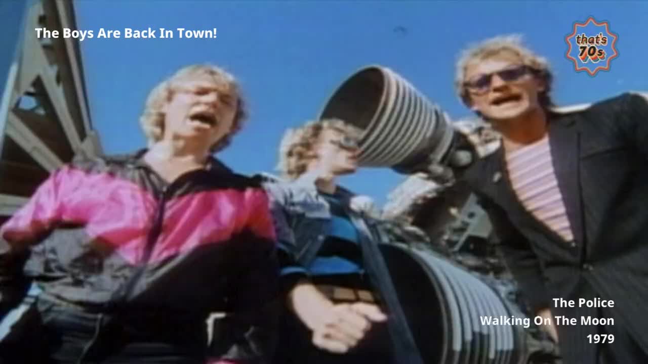Three blokes in 70s fashion are singing their hearts out, one in a bright pink and black jacket. They're standing near a large, metallic structure that looks like part of a rocket engine.