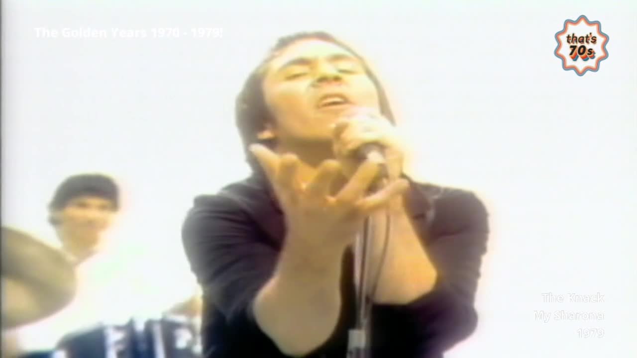 A singer belts into a microphone, his face tilted skyward and hands outstretched. Behind him, a drummer is barely visible, blurred against the bright backdrop.