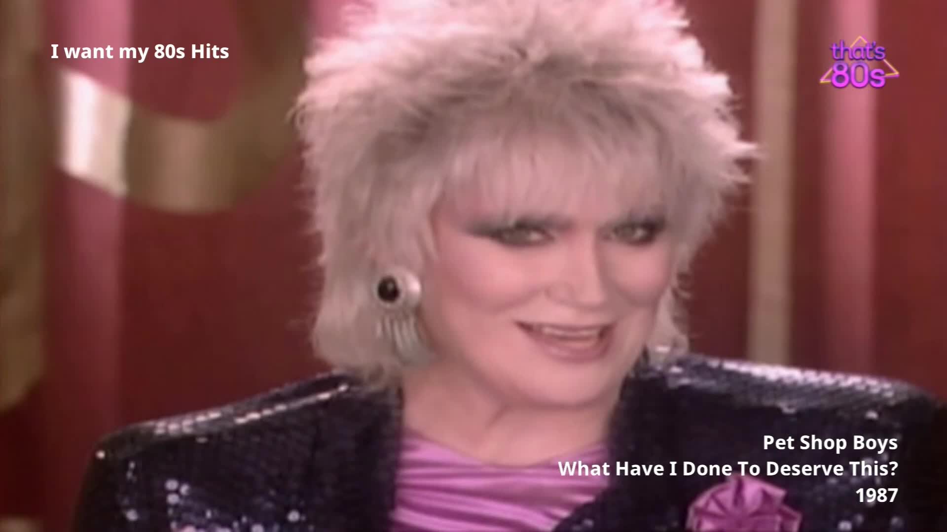 A woman with a voluminous, teased blonde hairstyle and dramatic eye makeup smiles faintly. She wears a sparkling black jacket over a shimmering pink top, her large, geometric earrings catching the light.