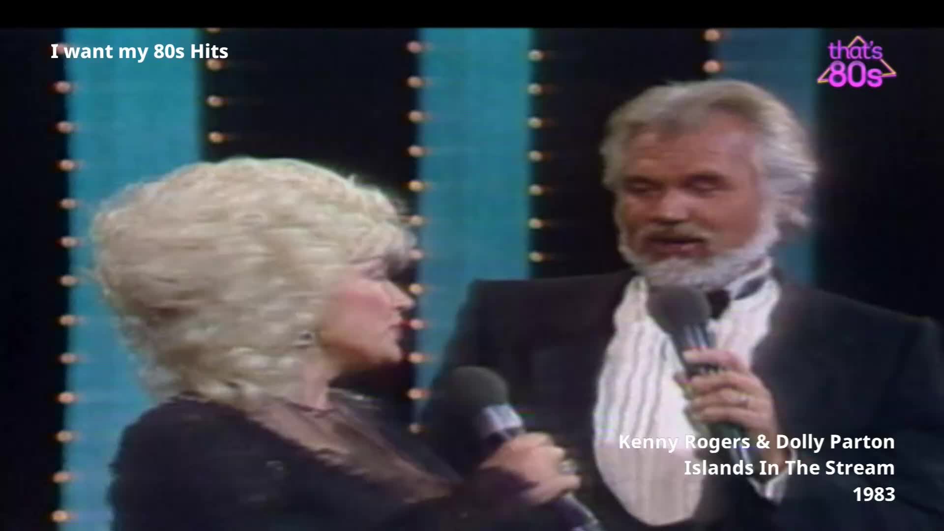 Dolly Parton, with her signature big blonde hair, stands close to Kenny Rogers as they both hold microphones. The stage lights behind them are arranged in vertical columns.