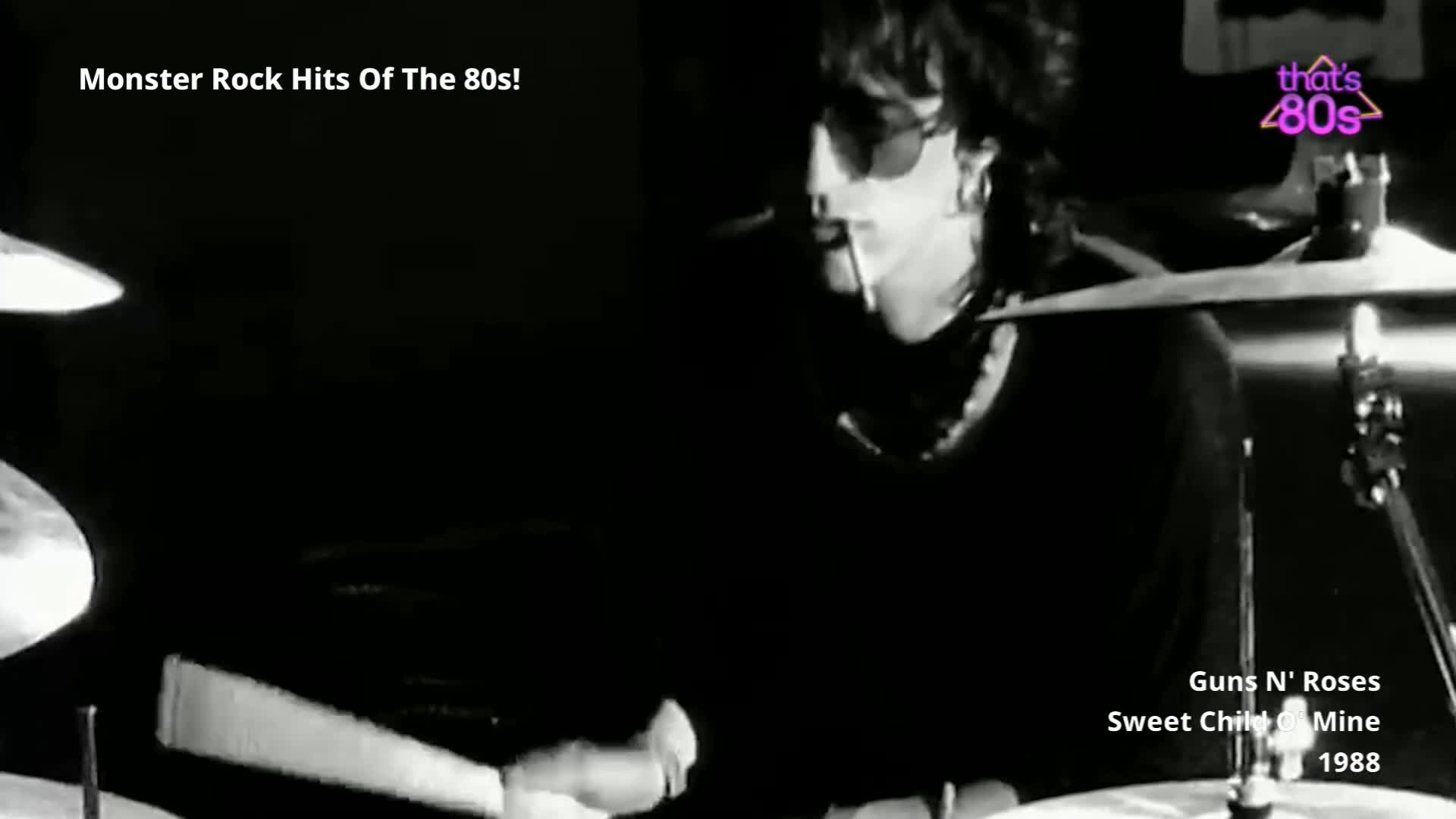 The drummer, wearing sunglasses, is hitting the cymbals with his sticks. His hair is dark and messy, and he's leaning into the beat.