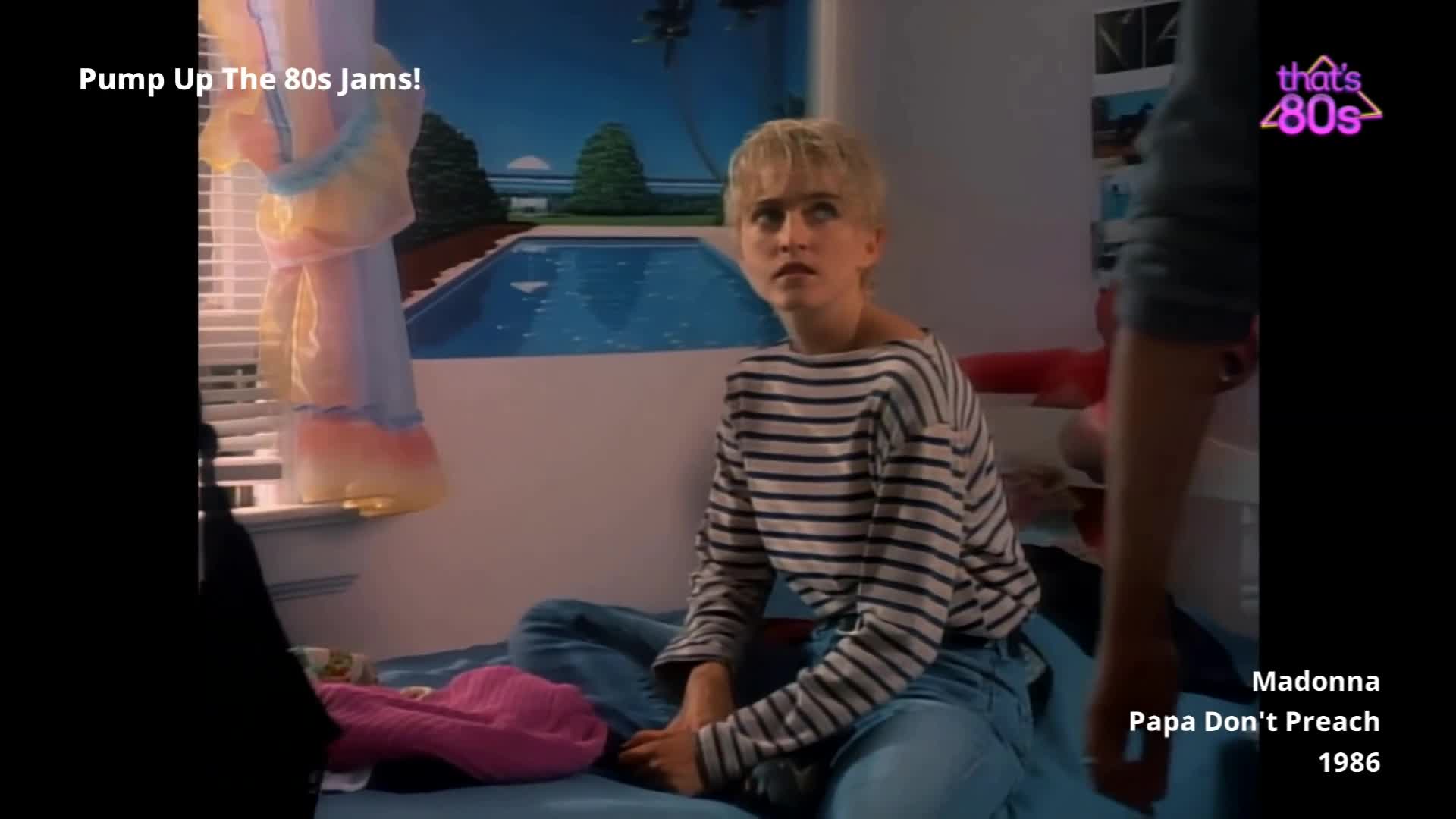 A young woman with short blonde hair, wearing a striped jumper and jeans, sits on a bed. She looks upwards with a worried expression, as another person's arm enters the frame from the right.