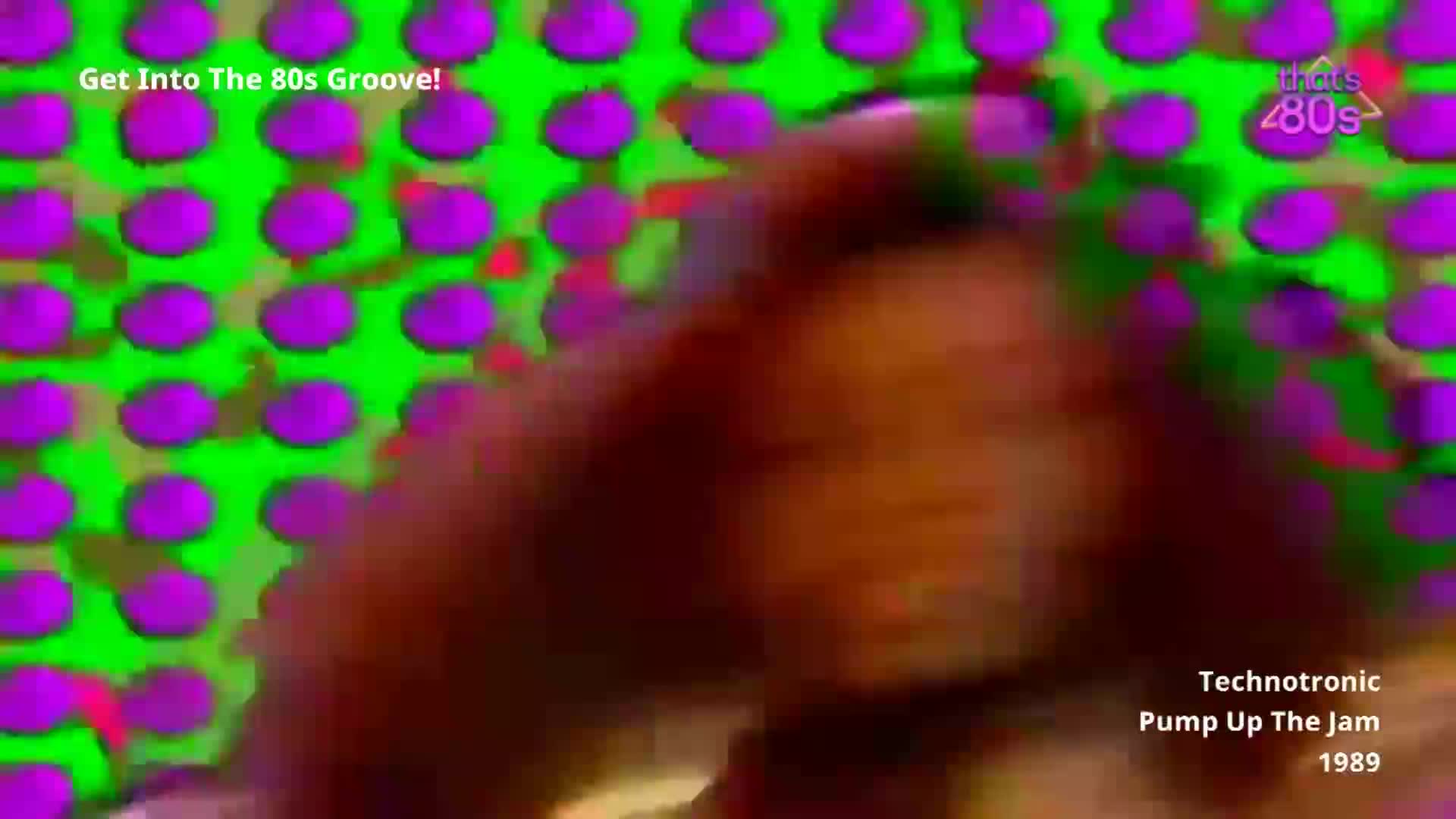 A woman with dark, curly hair is moving her head rapidly. The background is a dizzying pattern of bright green and purple circles.