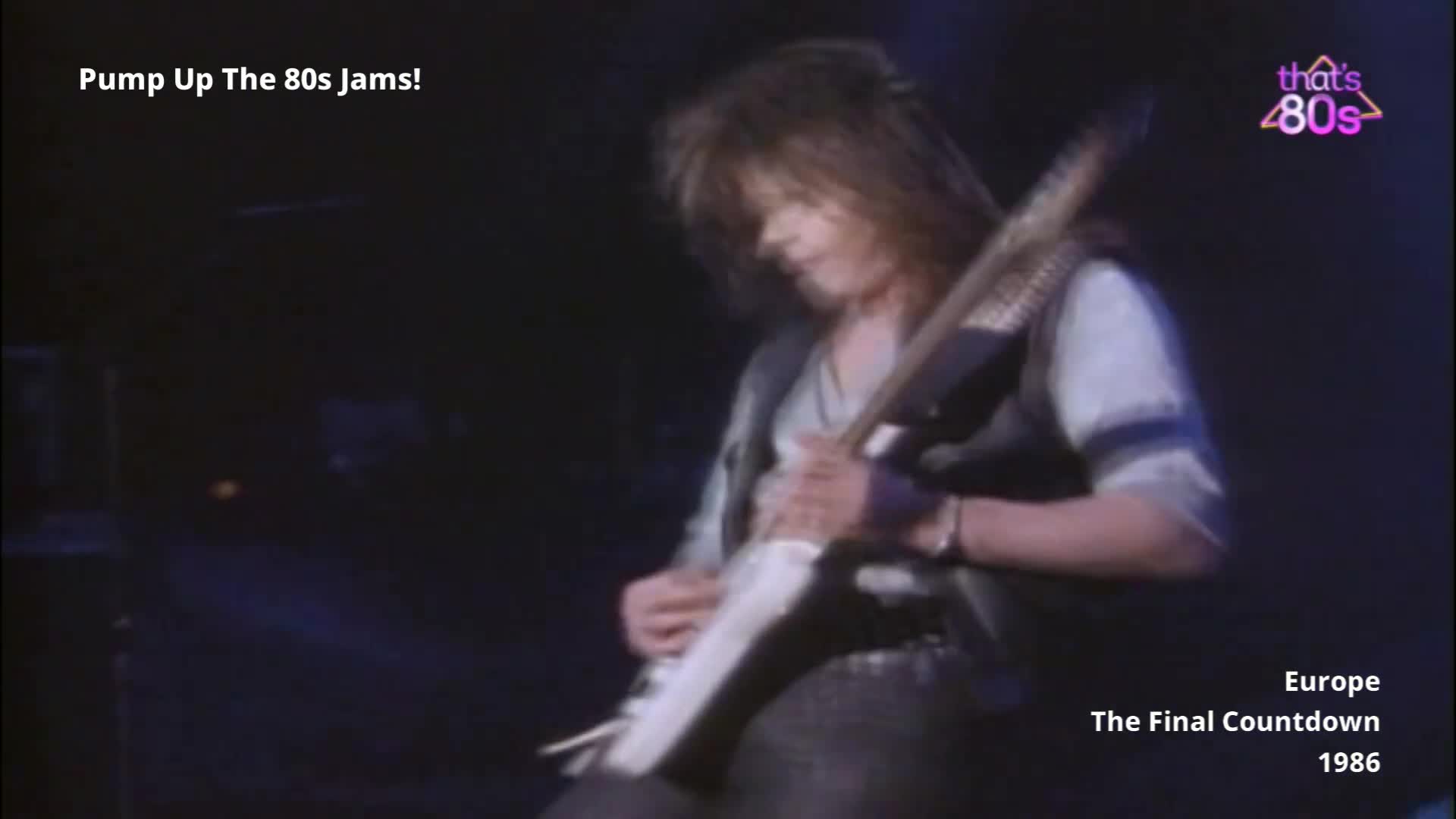 The guitarist's long hair flies as he rips into a solo on his white guitar. He's wearing a grey vest over a lighter shirt, and the crowd is a dark, indistinct mass in front of him.