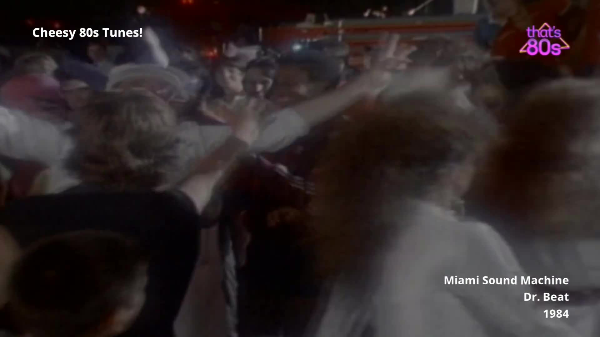 A crowd of people are dancing enthusiastically, their arms raised in the air. The lights above cast a warm glow on their faces as they move to the music.