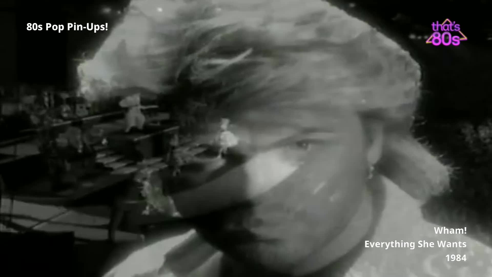 A young George Michael's face fills the frame, his hair a tousled mess. Behind him, a stage set with tiered platforms and figures in white outfits is visible.
