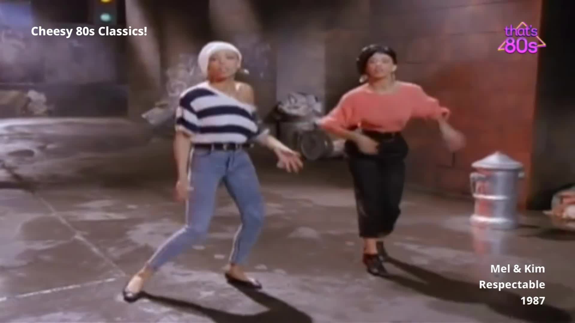 Two women are dancing in a warehouse-like space. The one on the left wears a striped off-the-shoulder top and jeans, while the other on the right is in a coral-coloured jumper and dark trousers.