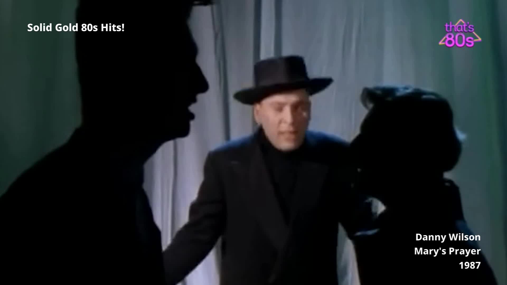 A man in a fedora and dark coat stands between two silhouetted figures. The backdrop is a pale curtain, and the lighting has a distinctly 80s feel.