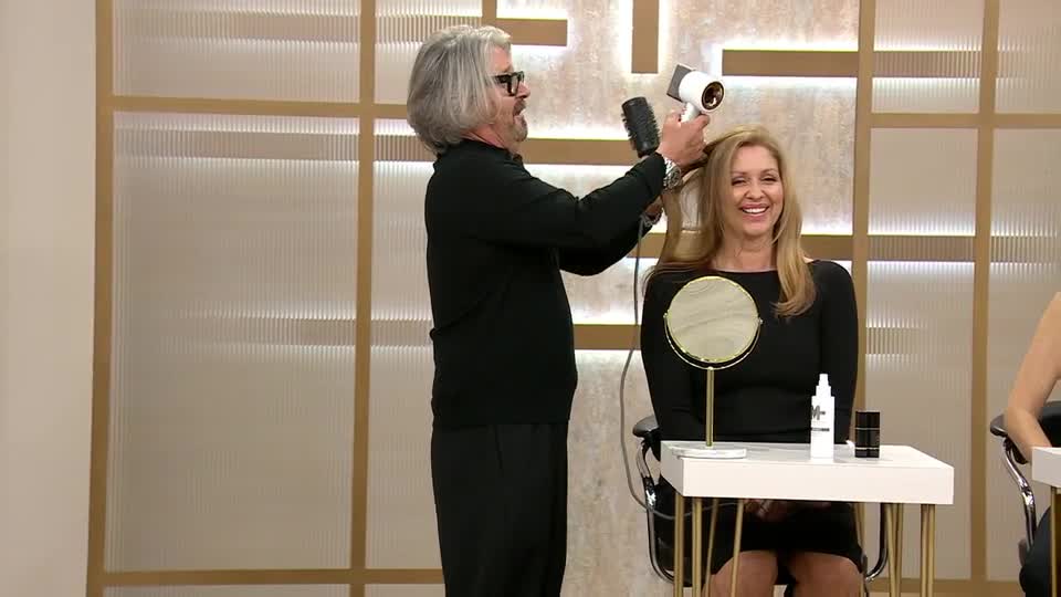 A man is carefully blow-drying a woman's hair, holding a brush in one hand and the dryer in the other. She beams as he works, a small mirror reflecting her happy expression.