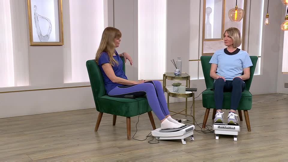 Two women sit in plush green chairs, each with their feet resting on a white vibrating platform. The woman on the left, wearing a blue top and leggings, watches her companion as the device beneath her feet hums. The woman on the right, in a lighter blue top and dark trousers, looks ahead, her trainers planted firmly on her own platform.