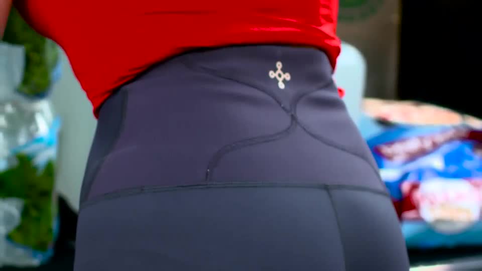 A woman in a red top and dark leggings turns, revealing a small, ornate logo on the back of her waistband. Behind her, bags of what look like fresh produce are stacked.